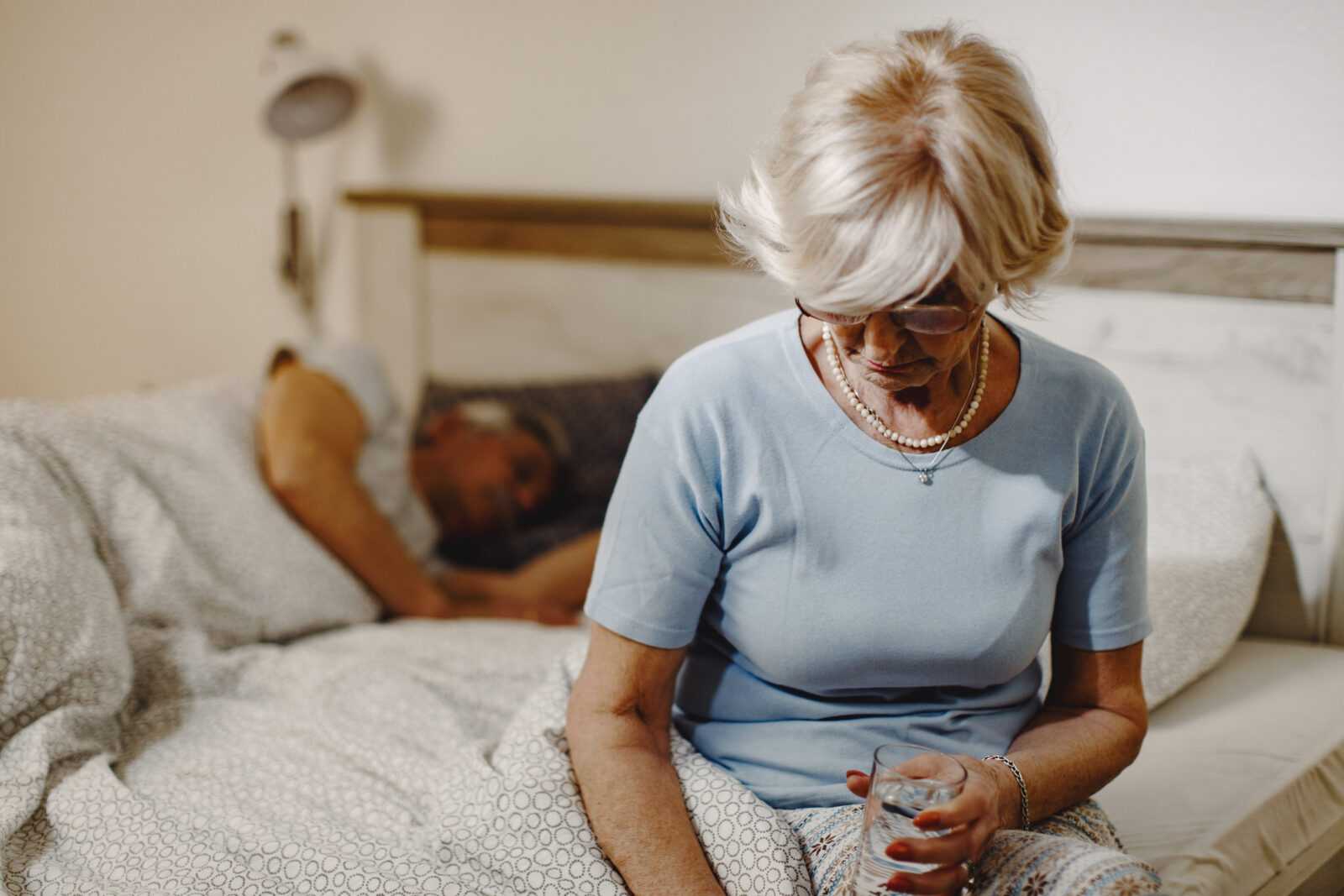 mature woman sitting bed holding glass water her husband is sleeping background